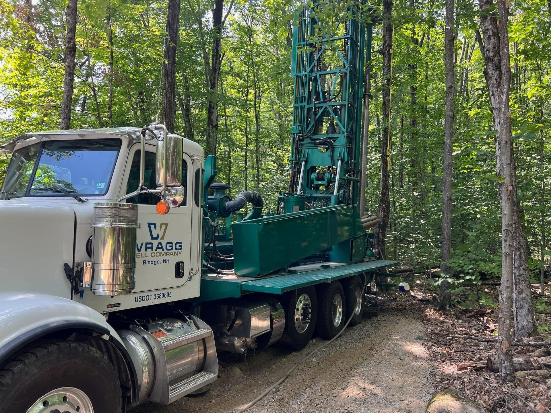 White truck carrying a green drilling rig on a forest dirt road