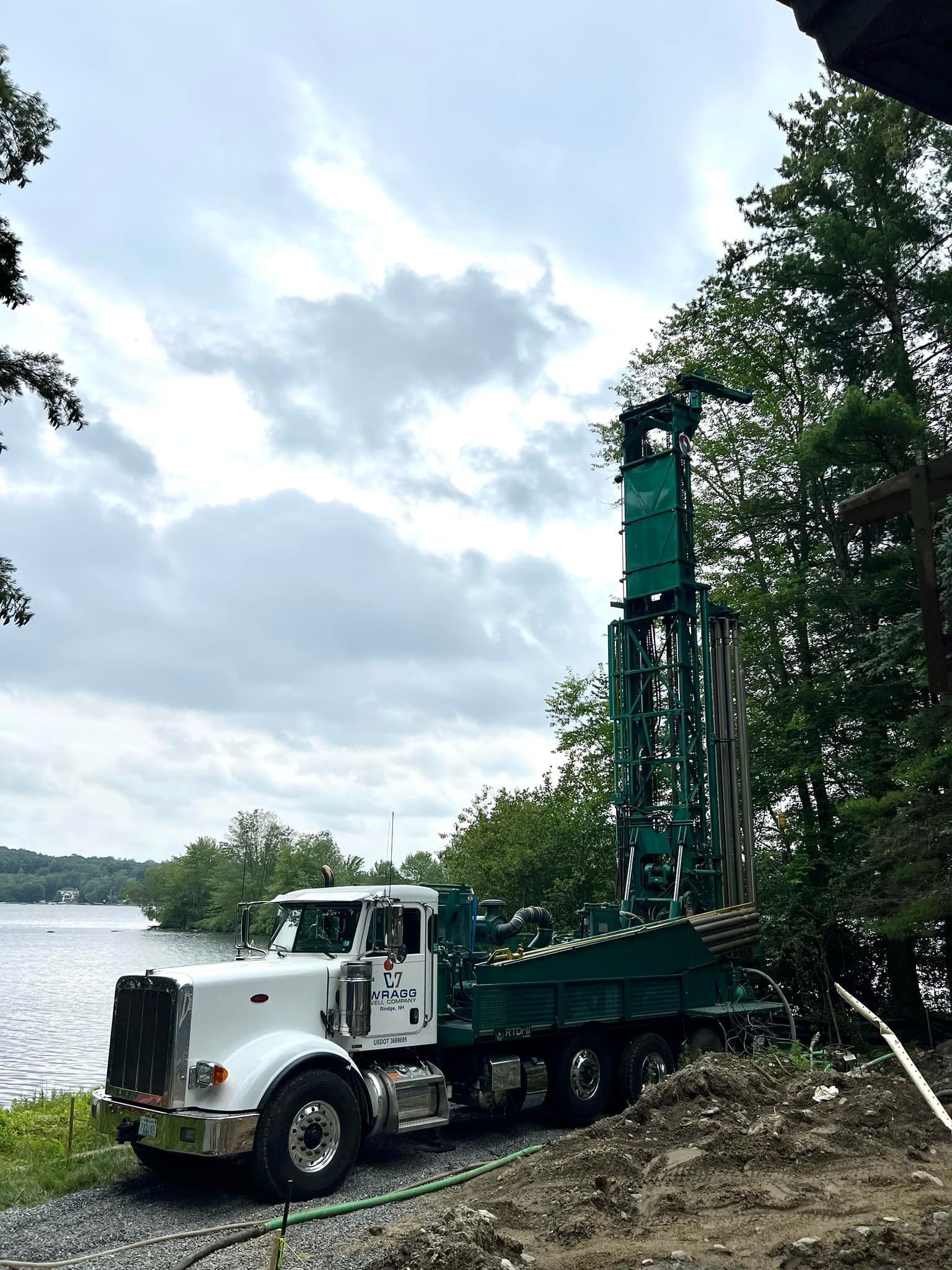 White flatbed drill truck parked by a lake with a tall green rig beside trees under cloudy sky