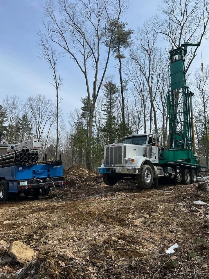 White truck beside green drilling rig in a wooded clearing with a blue equipment trailer