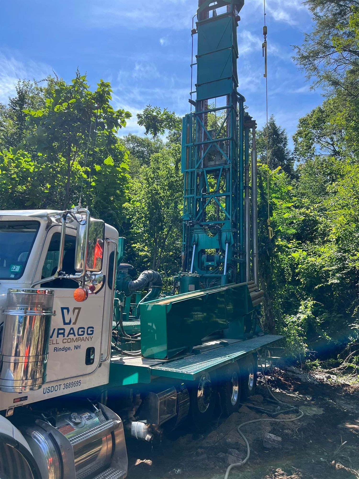 White crane truck with a tall blue drilling rig in a wooded area under a clear sky