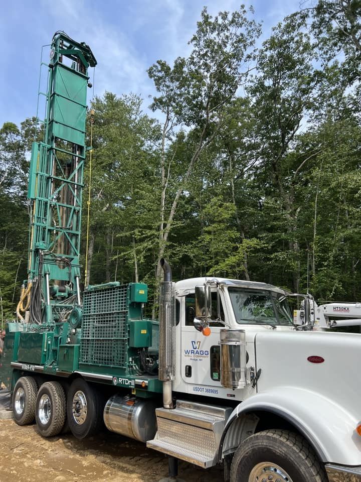 White utility truck with tall green drilling rig parked on a wooded roadside.