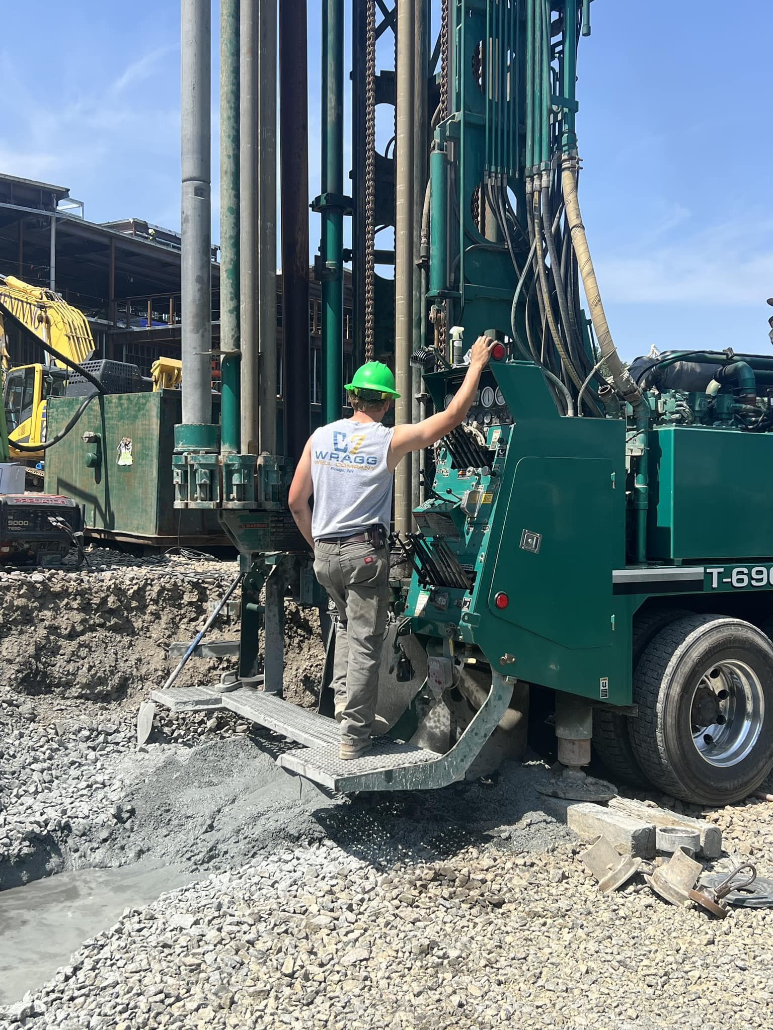 Worker in a hard hat stands beside a green drilling rig on a rocky construction site.
