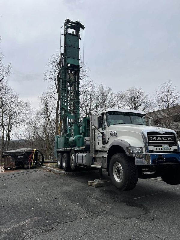 White Mack drilling truck with tall green rig parked on a paved lot under a gray sky