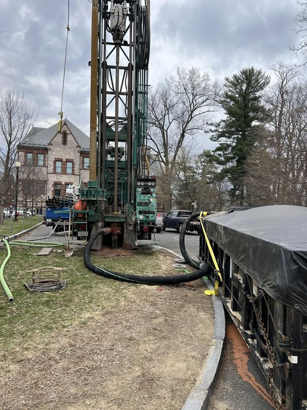 Drilling rig at a muddy construction site beside a covered trench and house in the background