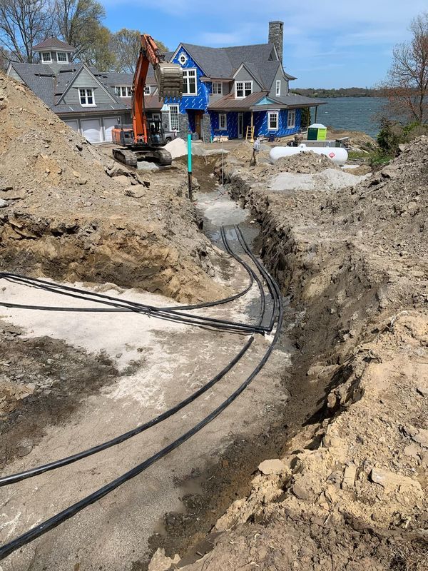 Excavator trenching beside a blue lakeside house, with pipes laid in a dirt worksite.