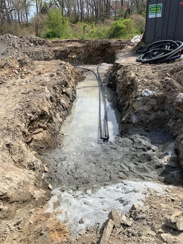 A muddy trench with standing water beside a black shed and coiled hoses in a wooded area.