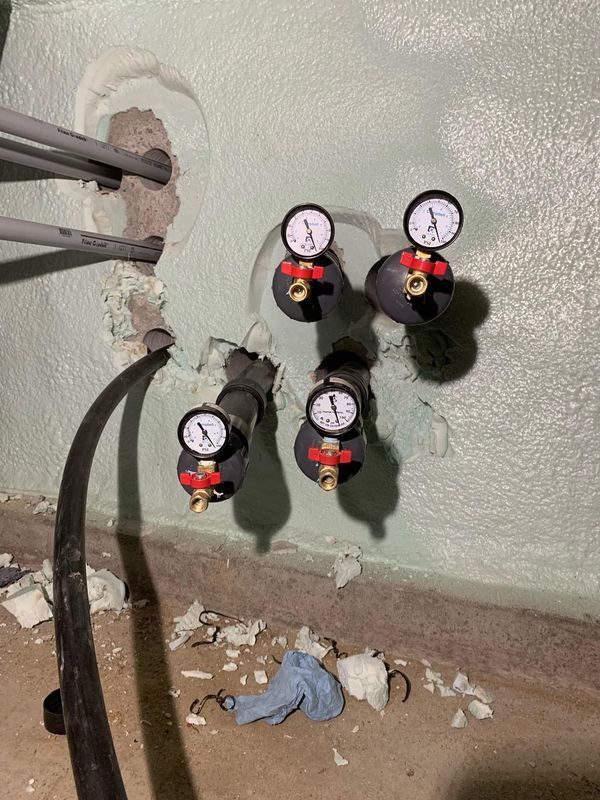 Four pressure gauges and valves on a wall-mounted pipe manifold near a rough hole in the wall