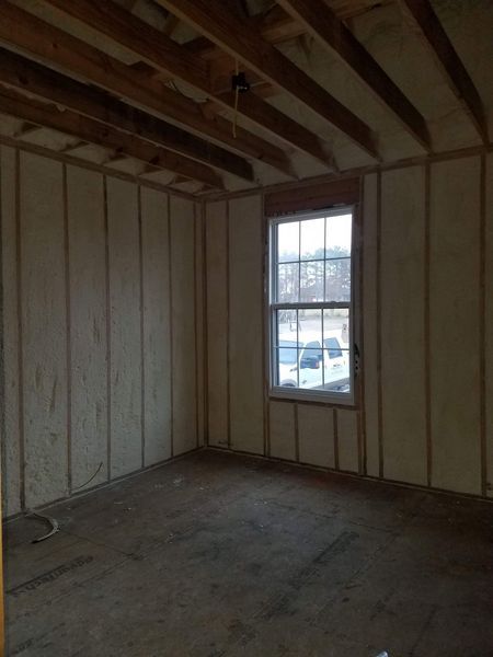 Empty room with spray foam insulation on walls, unfinished ceiling, and a window with outside view.