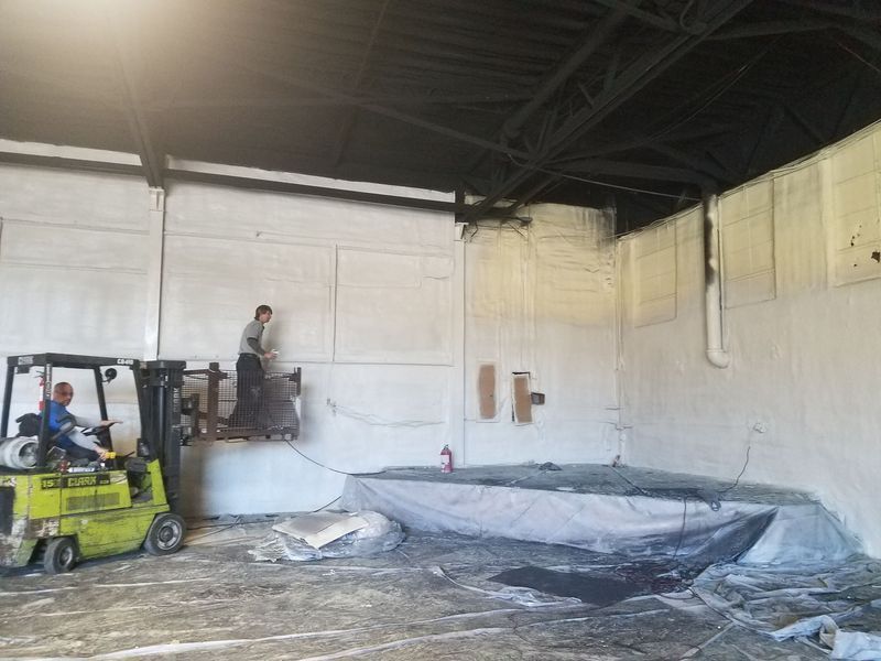 Workers on a forklift inside a building, spraying white material onto walls.