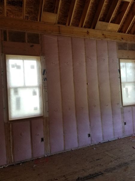 Interior view of a room under construction; pink insulation between wood studs, two windows, and unfinished wood flooring.