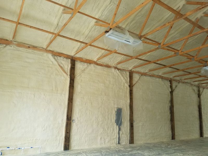 Interior of a building insulated with light tan spray foam. Wooden beams and concrete floor visible.