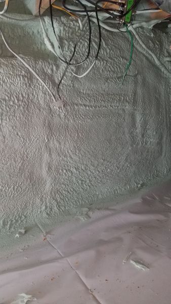 White spray foam insulation on a basement wall, with visible electrical wires and a protective plastic sheet.