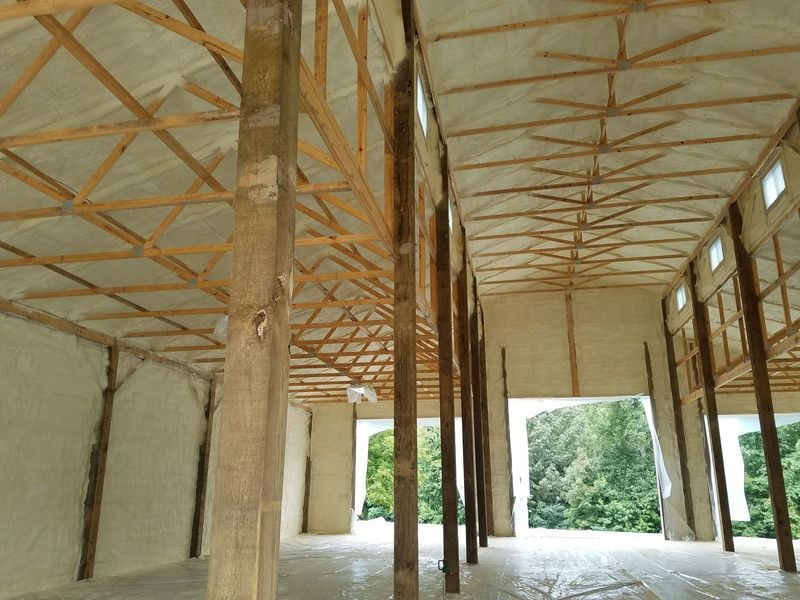 Interior view of a barn with exposed wooden beams, light-colored insulation, and an open doorway to a green landscape.
