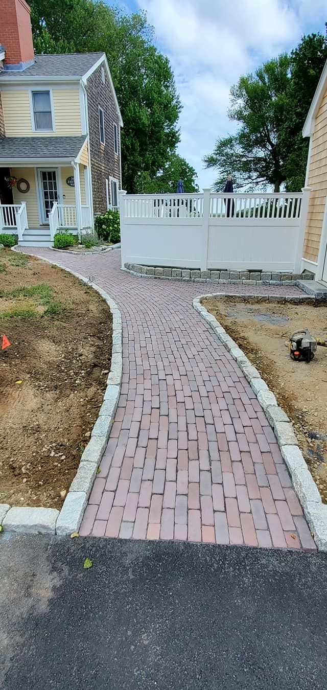 A brick walkway leading to a house with a white fence.
