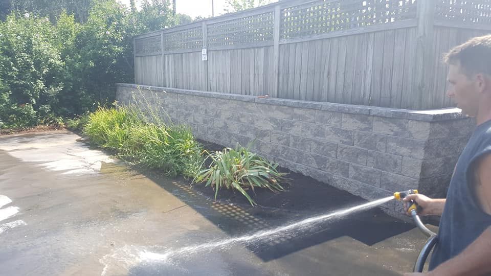 A man is watering plants with a hose in front of a fence.