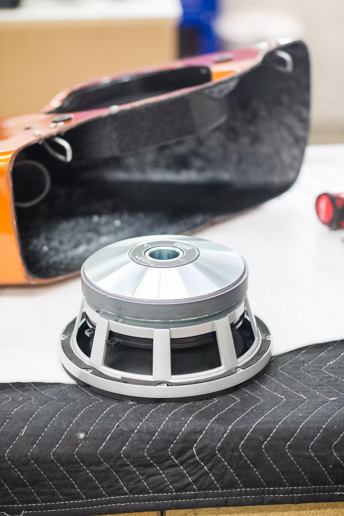 Silver speaker component on a dark quilted fabric, with an orange case in the background