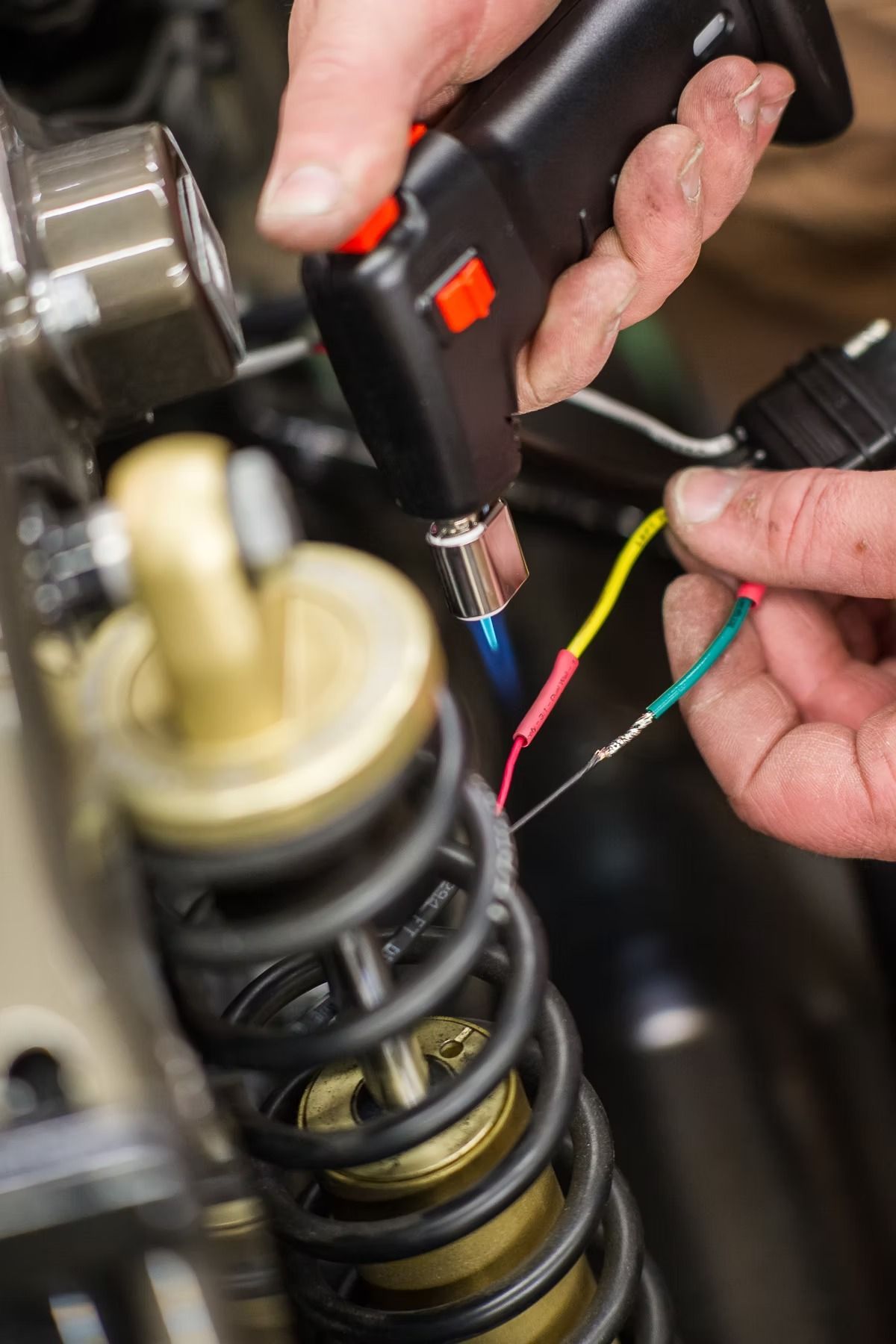 Hands using a soldering iron on colored wires near a car suspension spring.