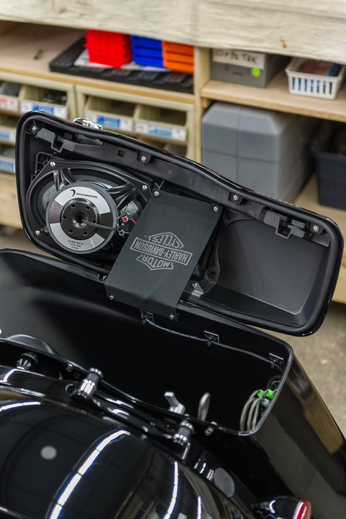 Open motorcycle saddlebag with visible speaker and wiring, glossy black bike in a garage.