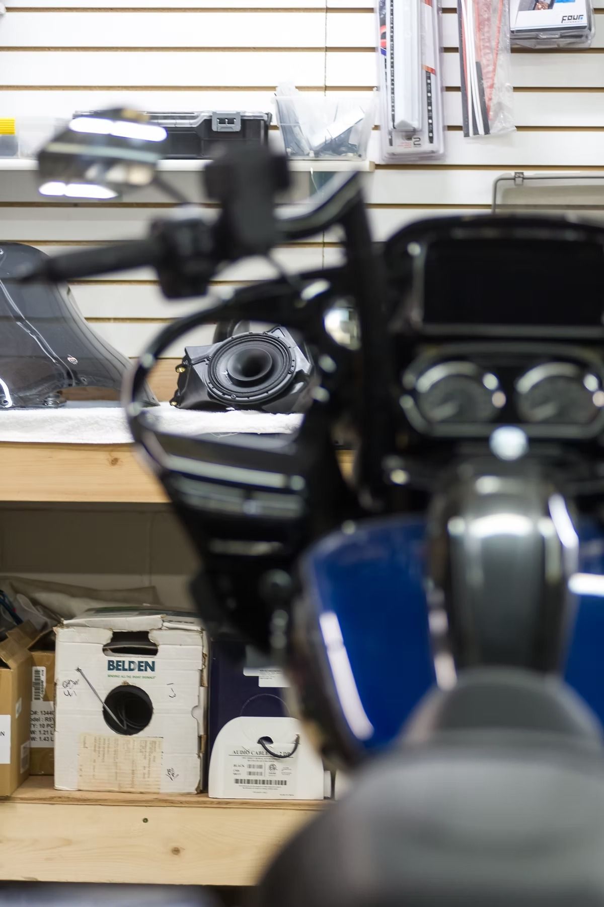 A blue scooter in a shop, viewed from the front with handlebars and dashboard visible.