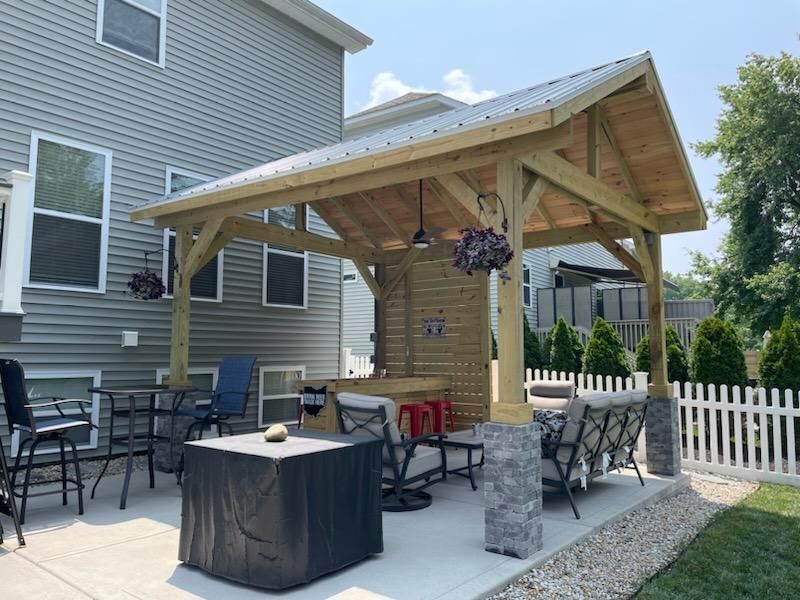 A wooden pavilion with a metal roof is in the backyard of a house.