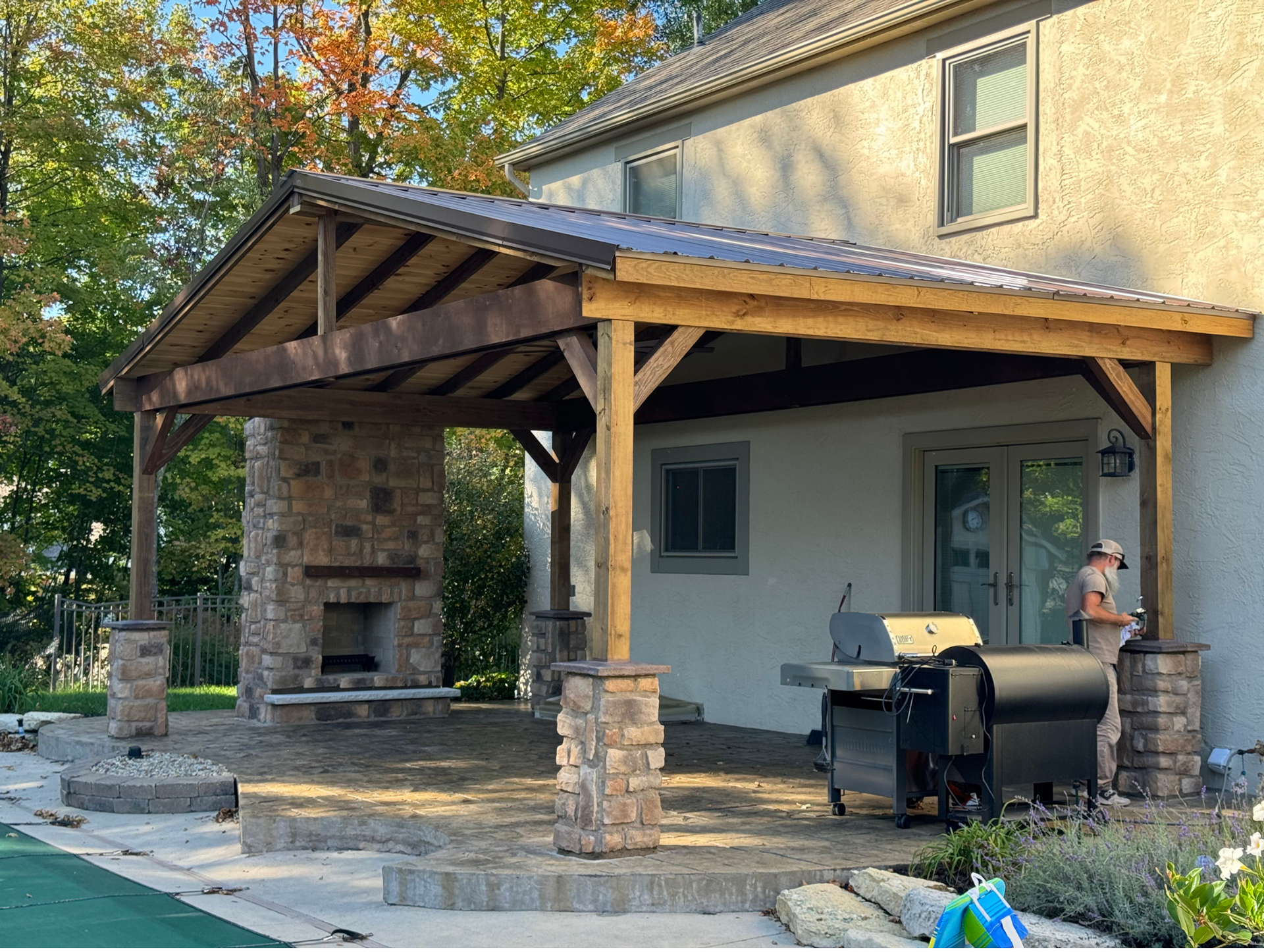 A man is standing under a wooden covered patio with a grill and fireplace.
