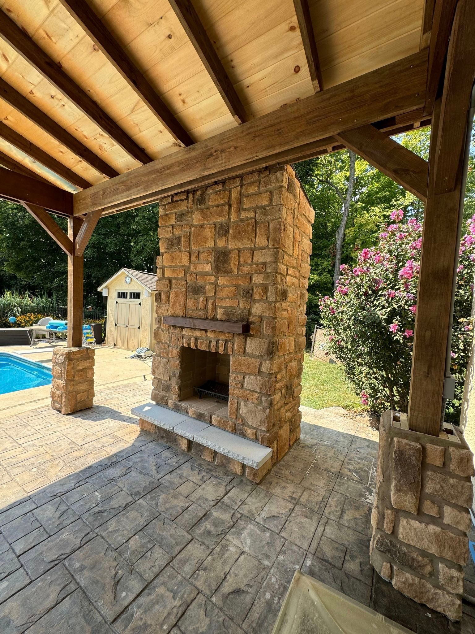 A stone fireplace under a wooden roof next to a pool.