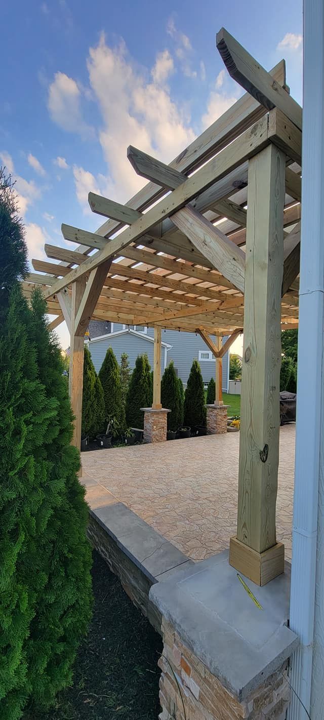 A wooden pergola is sitting on top of a gravel driveway next to a house.