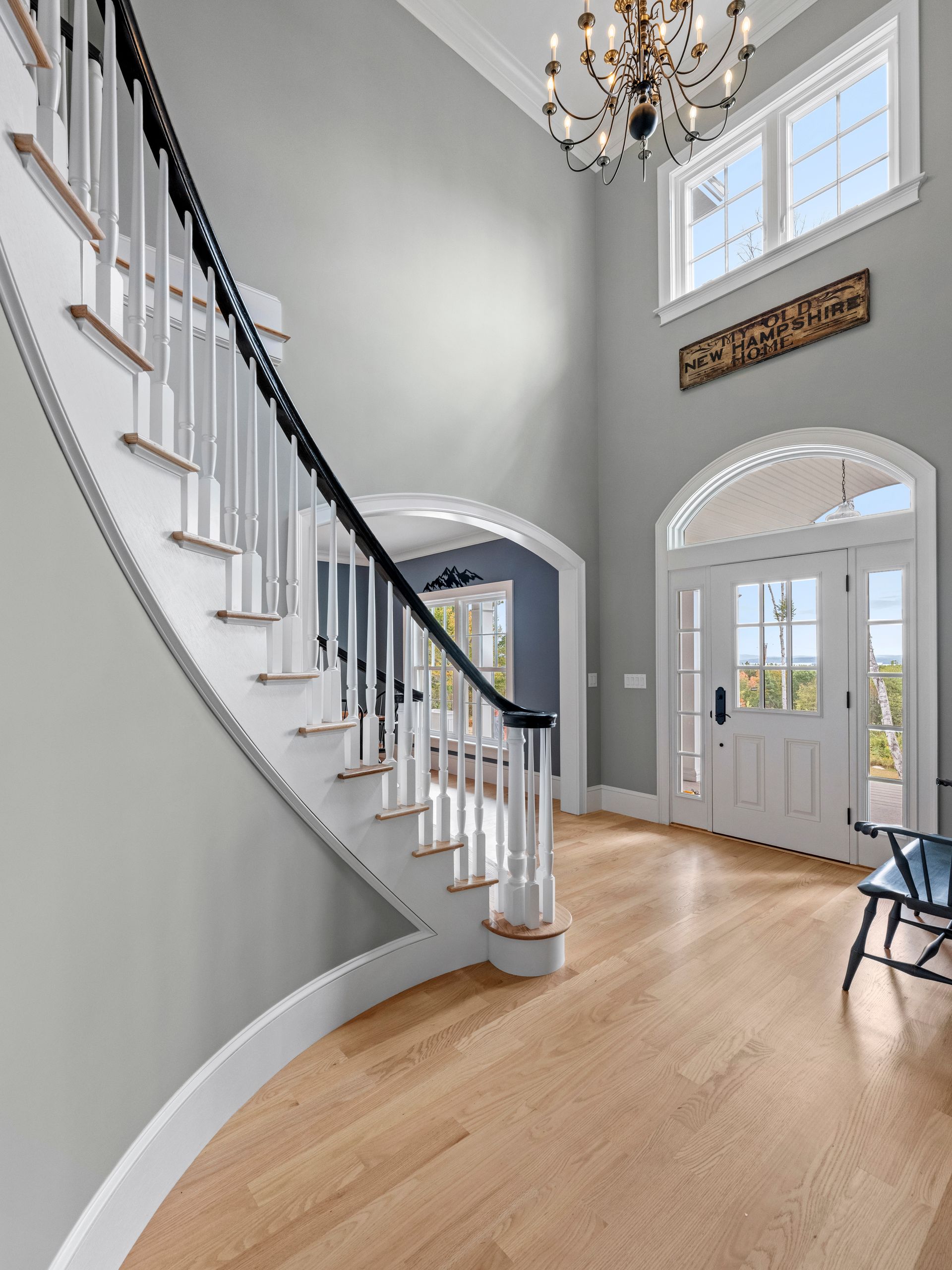 Grand entrance hall with curved staircase, hardwood floors, and gray walls. White trim, black railing, and chandelier.