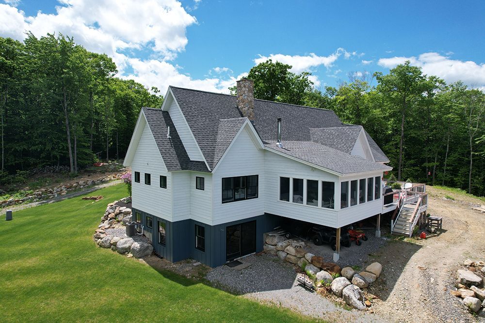 White house with dark blue trim, gray roof, surrounded by trees and rocks, on a sunny day.