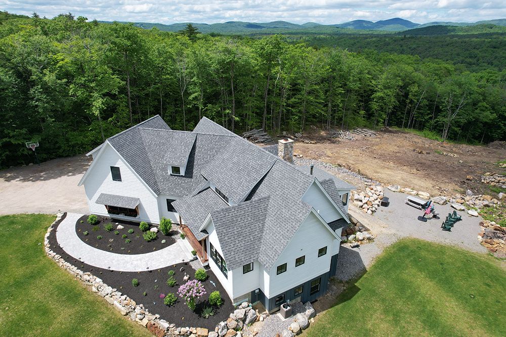 White house with gray roof, surrounded by green lawn and trees, mountains in background.