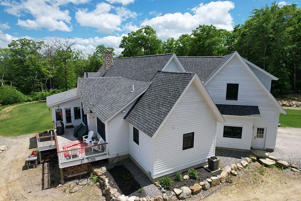 White house with gray roof, wooden deck, surrounded by trees under a blue sky.