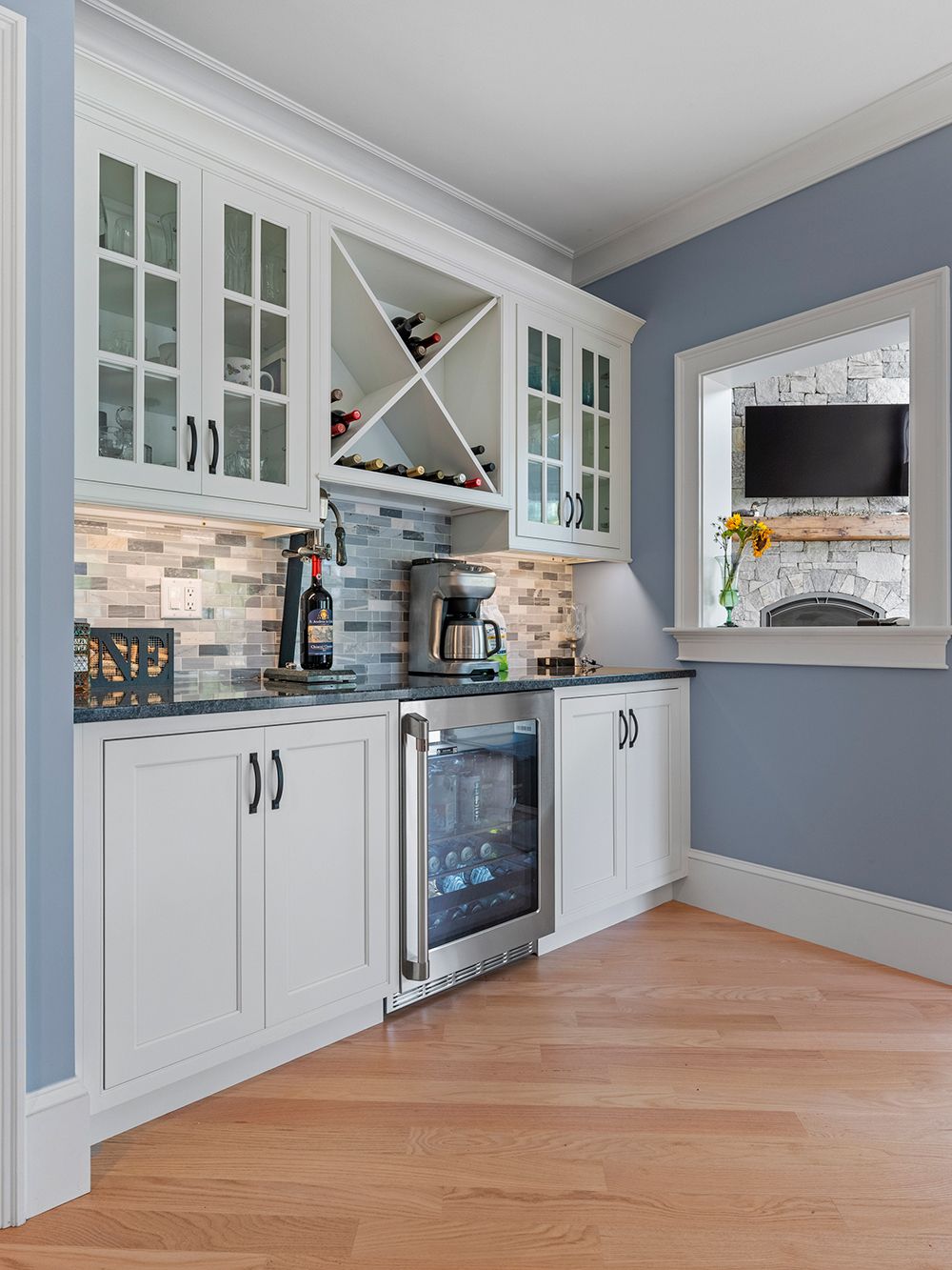 White cabinetry with a wine rack, mini-fridge, and built-in coffee maker in a light-blue room, wooden floor.