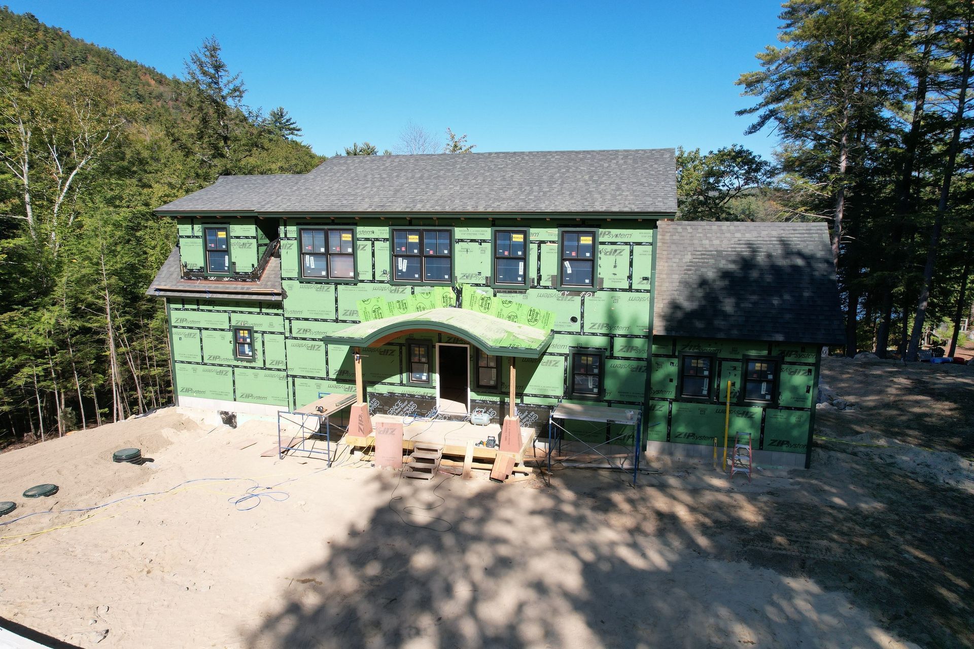 Two-story house under construction, green sheathing, dark roof, windows, porch, surrounded by trees and dirt.
