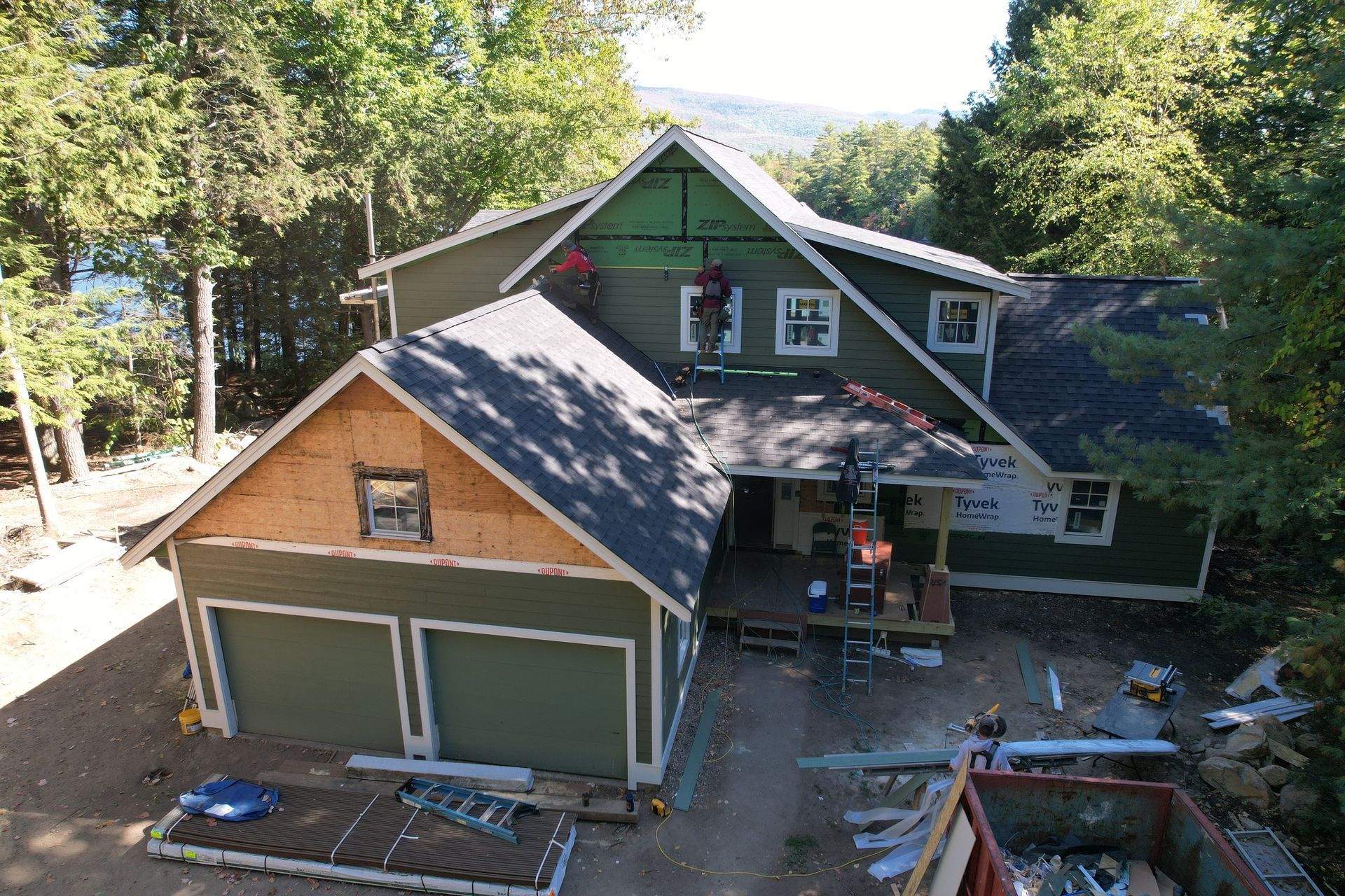 House under construction with green siding, workers on roof, surrounded by trees.
