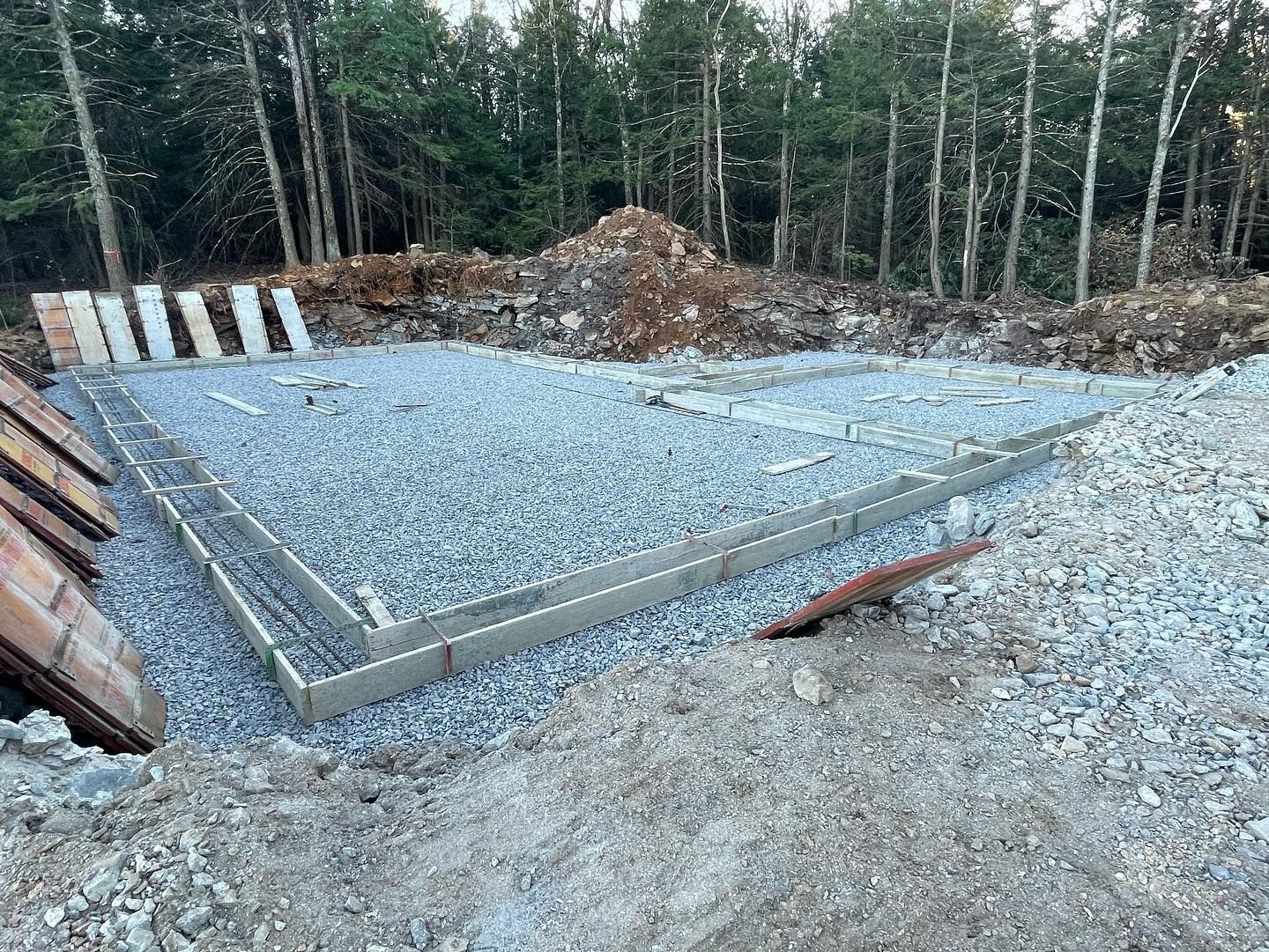 Foundation of a building, framed in concrete, filled with gravel, in a wooded area.