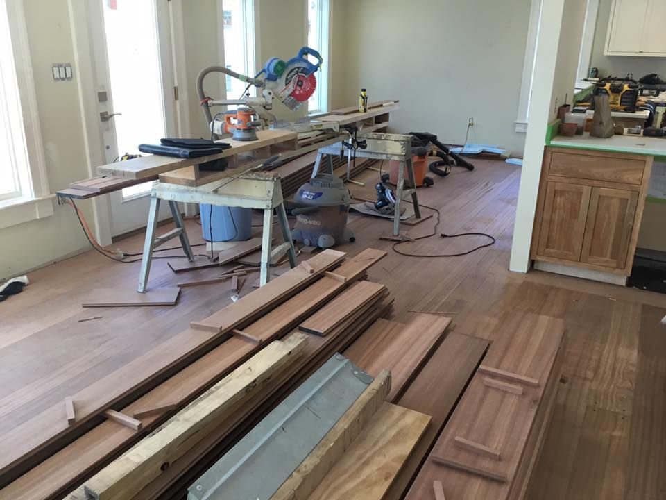 A room with hardwood flooring under construction. A sawhorse supports a miter saw. Wood planks are stacked nearby.