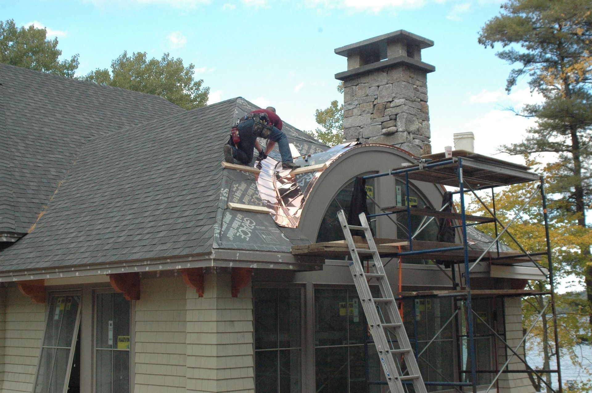Workers on a roof with scaffolding, replacing shingles near a stone chimney.