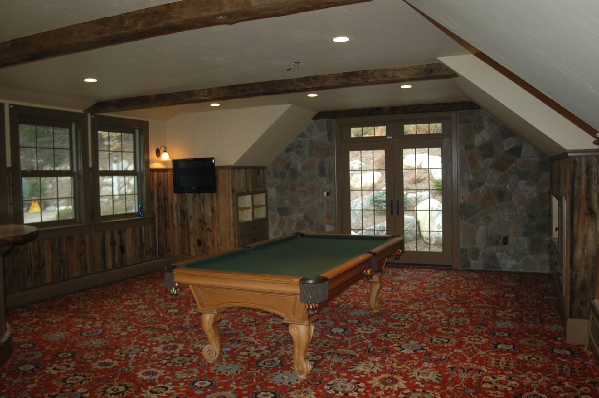 Pool table in a rustic room with stone wall, wooden beams, windows, TV, and red patterned rug.