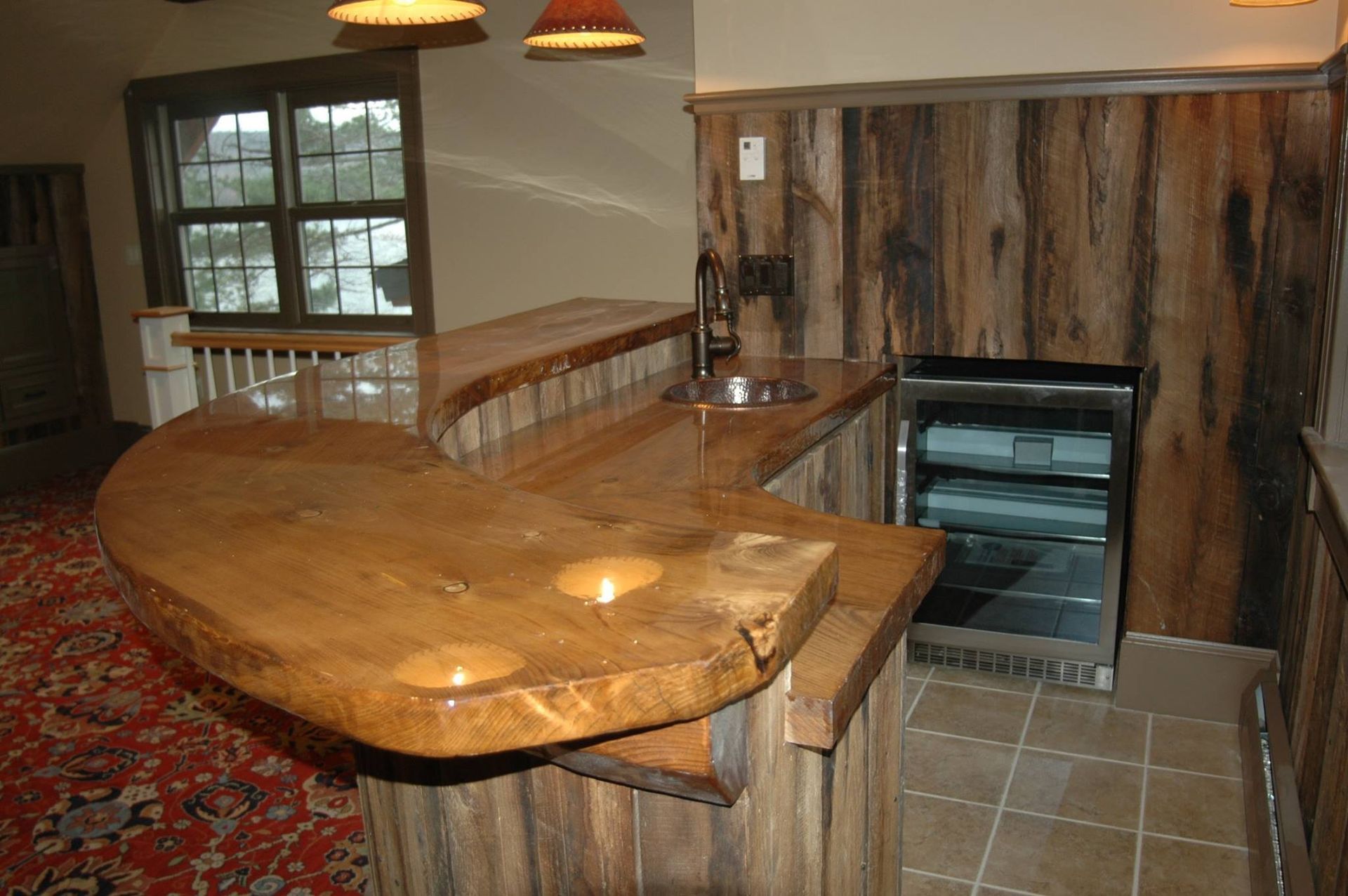 Rustic wooden bar with built-in mini fridge and sink, window in the background.