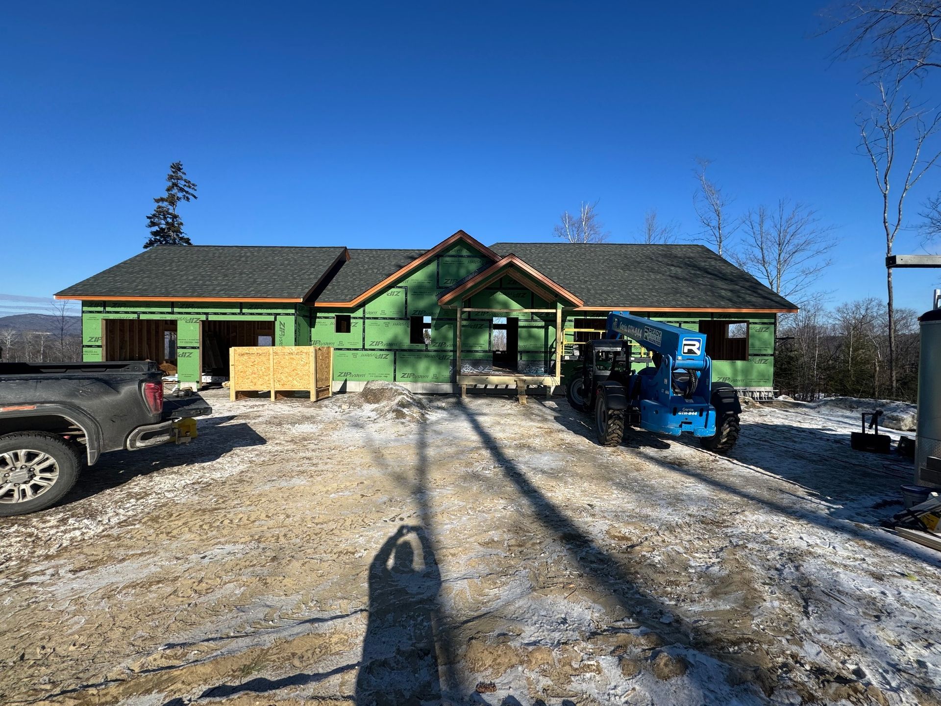 Construction of a single-story house on a snowy lot; green sheathing, blue lift, truck, clear sky.