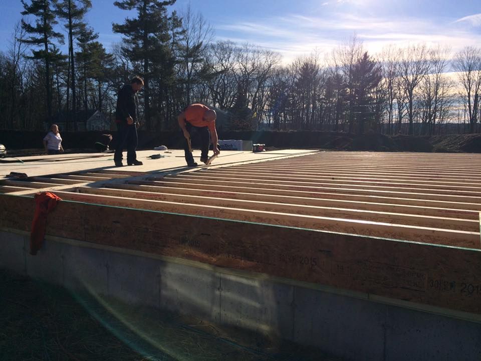 Construction workers on a wooden floor foundation, outdoors with trees in the background, sunny day.