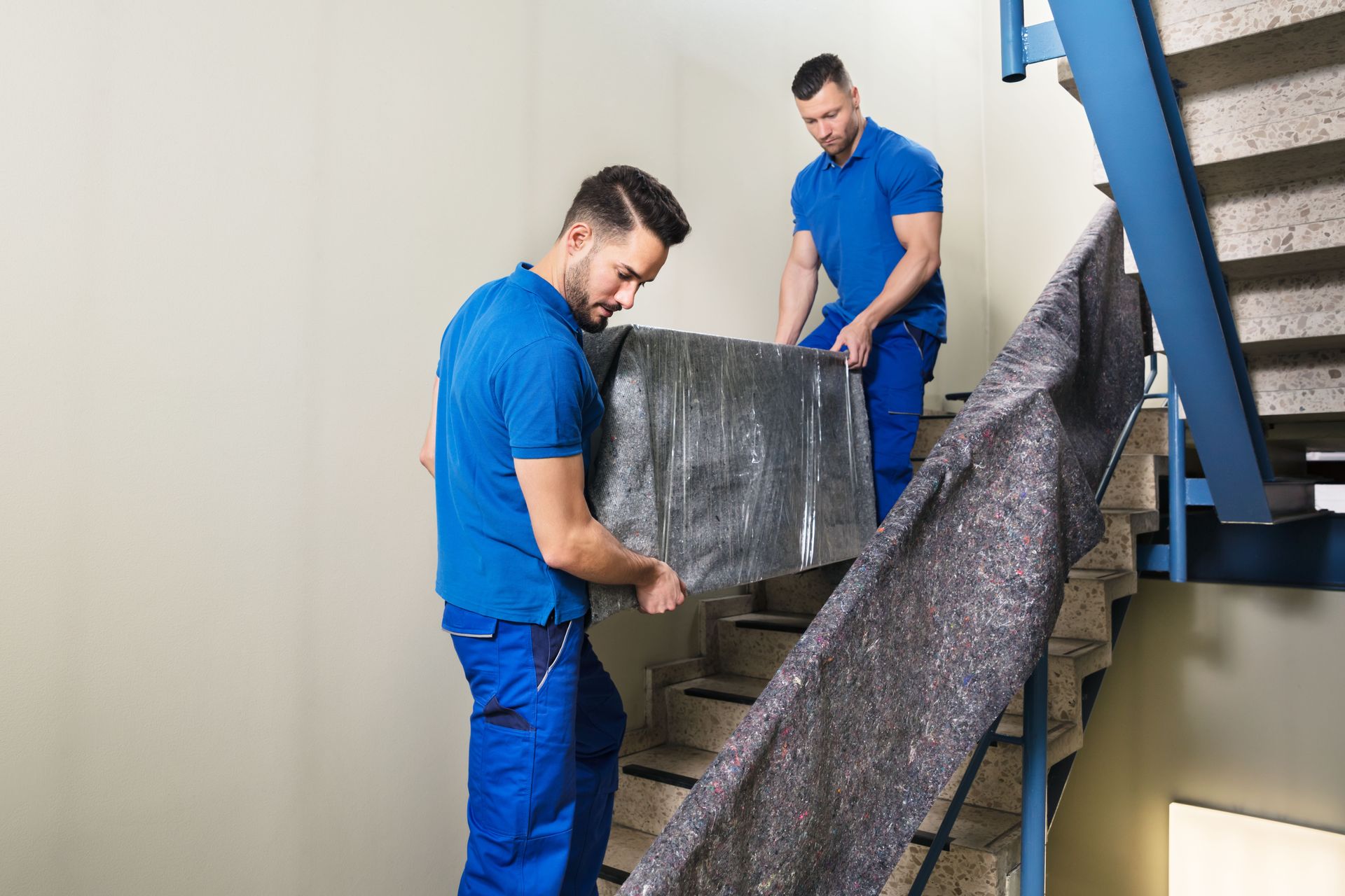 Two movers in blue uniforms carry a wrapped piece of furniture down an indoor staircase.