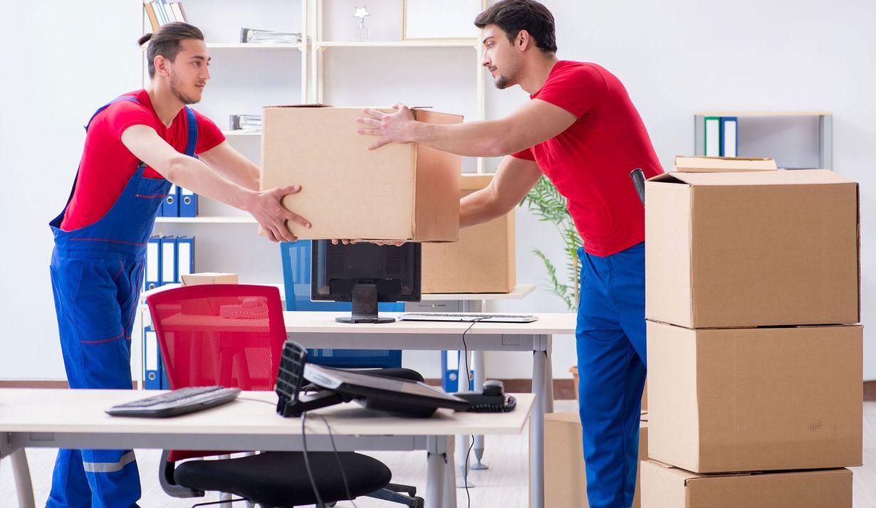Two movers in red shirts and blue overalls work together to carry a cardboard box in an office setting.