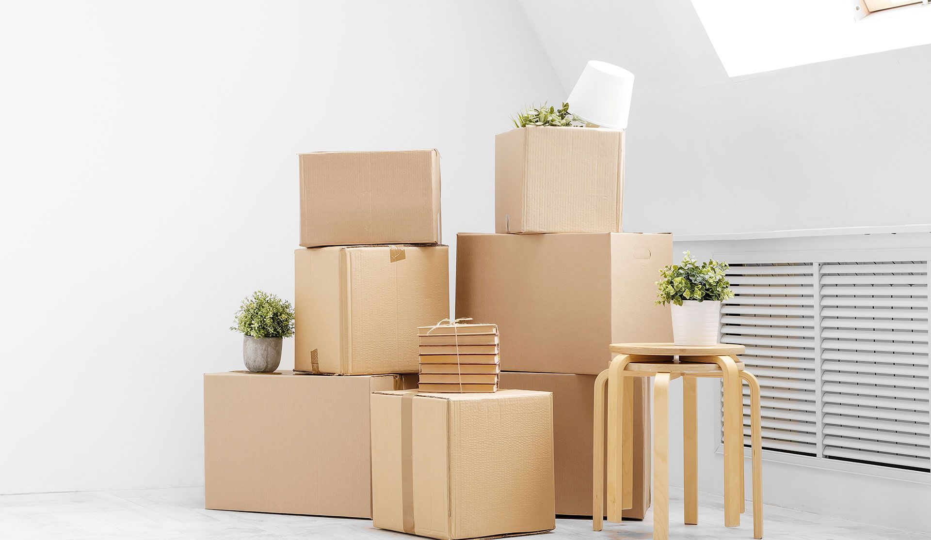 A stack of brown cardboard moving boxes arranged in a bright room with two small potted plants and a wooden stool.