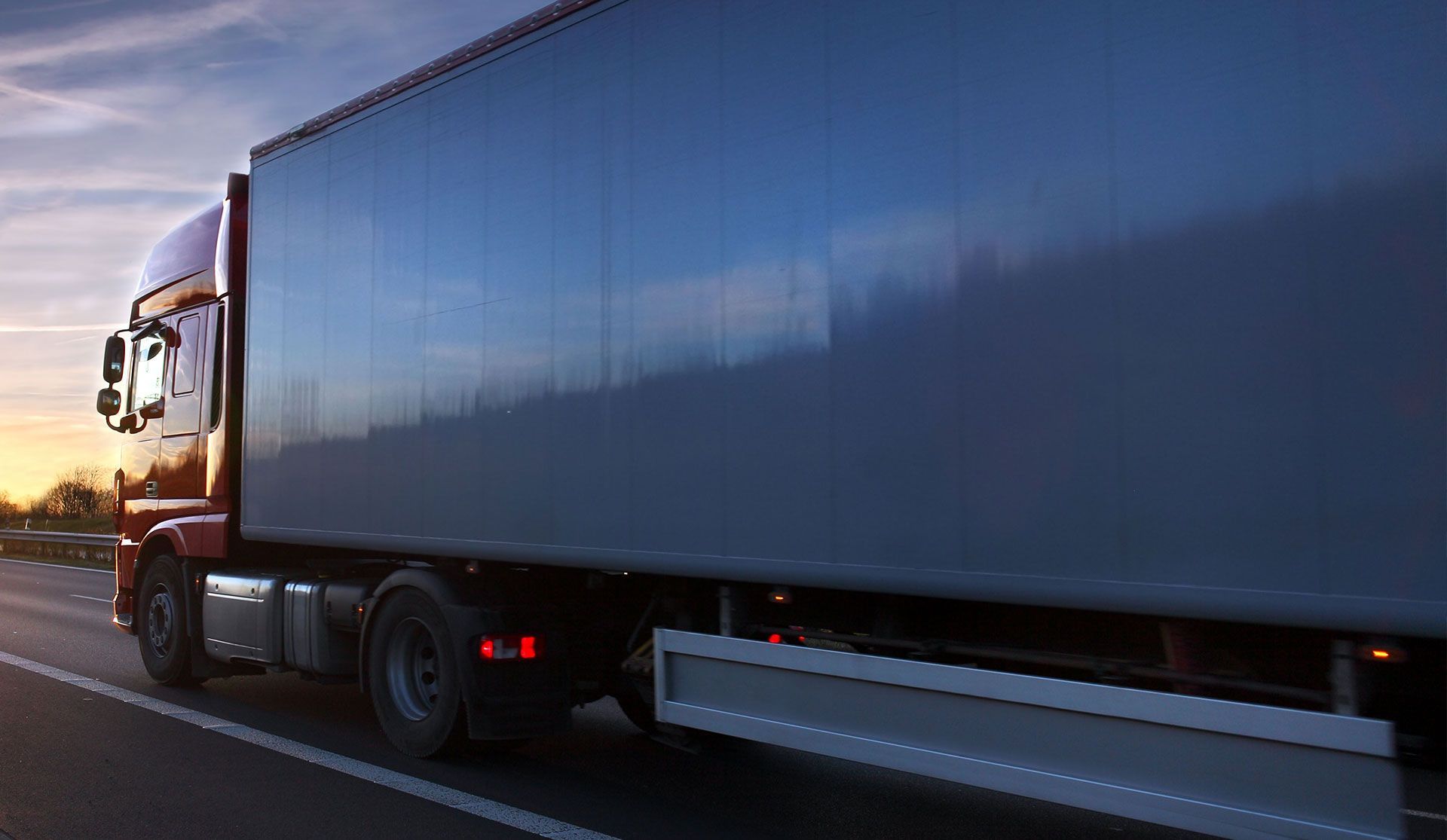A red semi-truck driving on a road at sunset, viewed from the side.