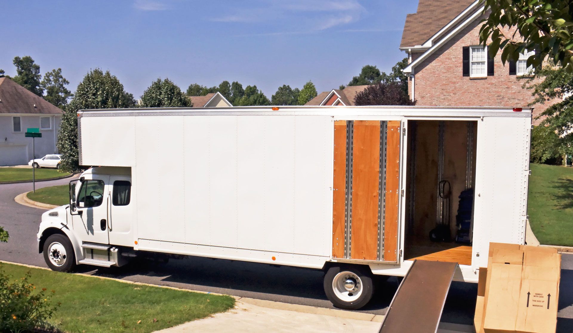 A white moving truck parked on a suburban street with its side door open and a ramp extended to the ground.