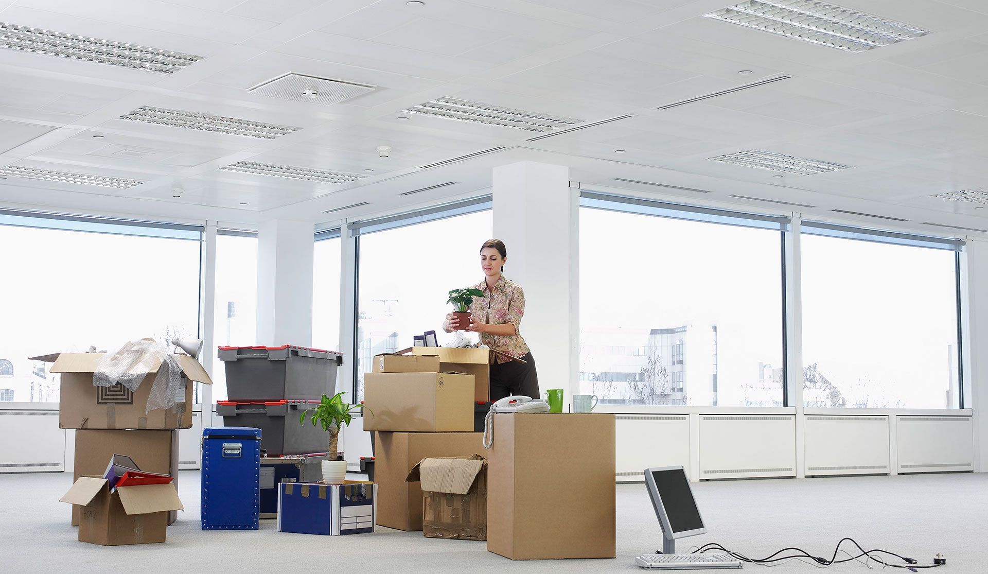 A person packs belongings into cardboard boxes and plastic bins in an empty office with large, bright windows.