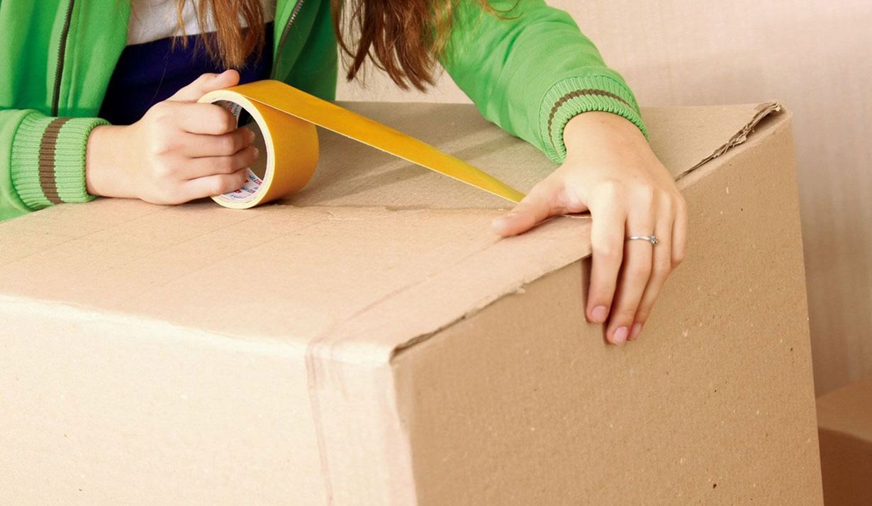 A person wearing a green jacket seals a cardboard shipping box with a roll of tan packing tape.
