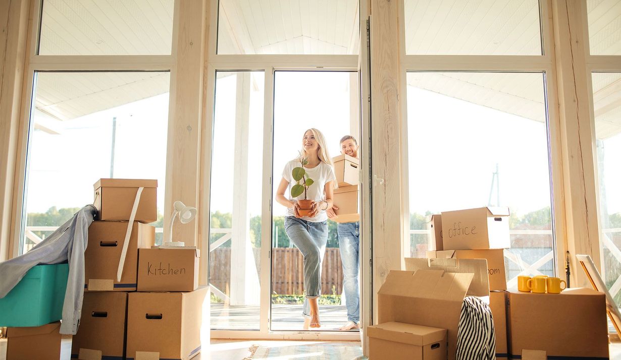 A couple carrying boxes and a potted plant enters a sunlit, unfurnished house filled with cardboard moving boxes.