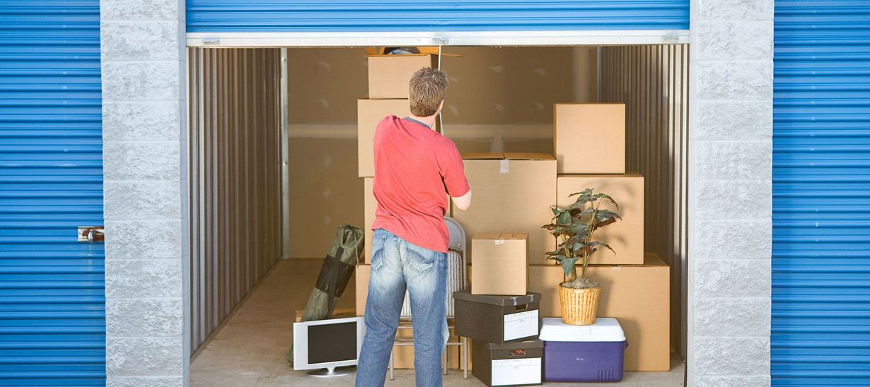 A person in a red shirt stands inside a storage unit filled with various cardboard moving boxes and a potted plant.